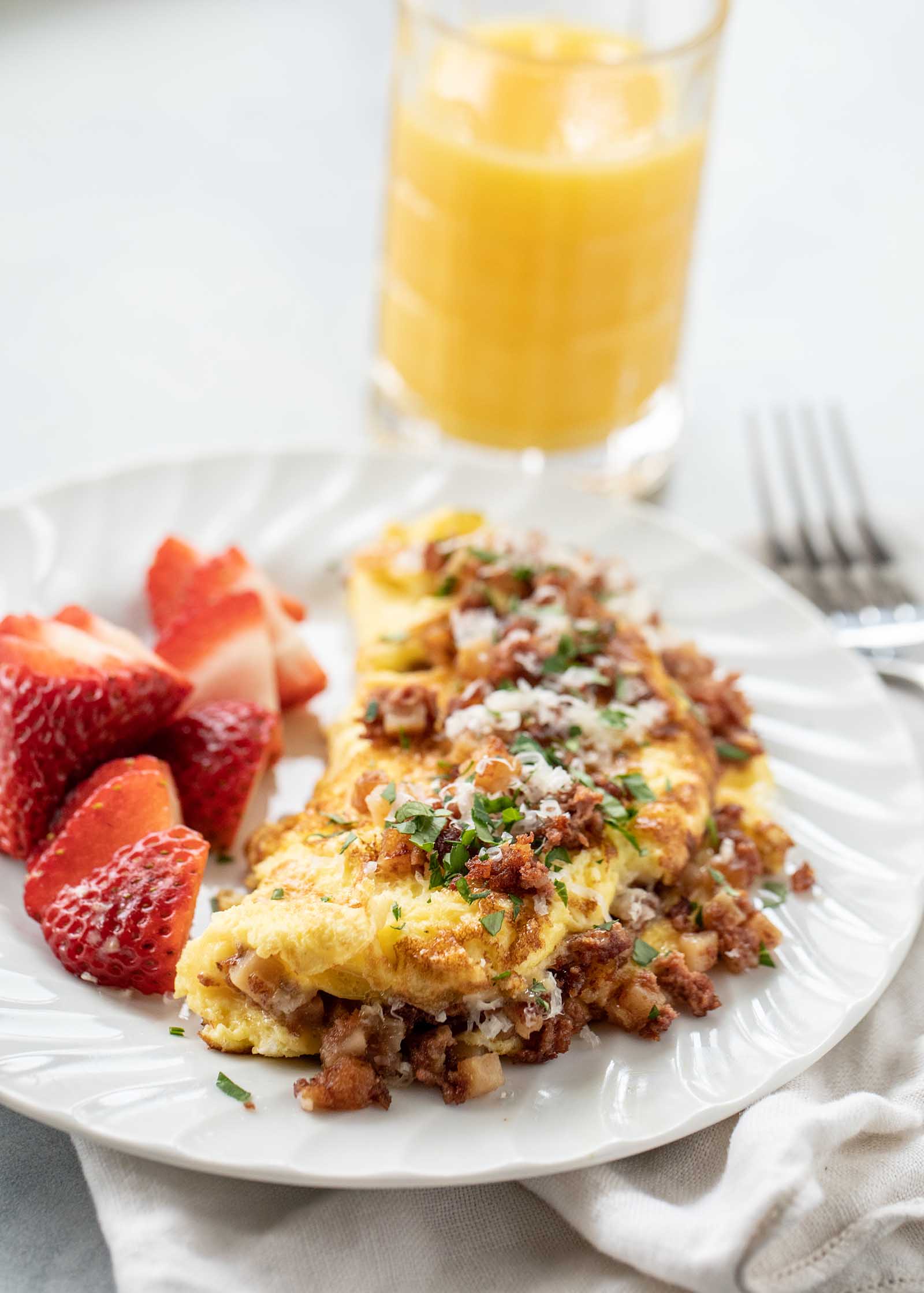 The perfect homemade corned beef omelet is on a white plate. Browned corned beef, grated parmesan cheese and fresh parsely are on top and to the sides of the omelet.Halved strawberries are on the left of the omelet. A fork and glass of orange juice are above the plate and to the right.
