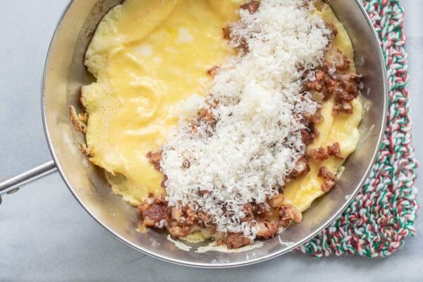 A skillet is on a green and red striped pot holder. The skillet has cooked eggs in the bottom of the pan and the chopped corned beef and shredded parmesan on one side of the cooked eggs..