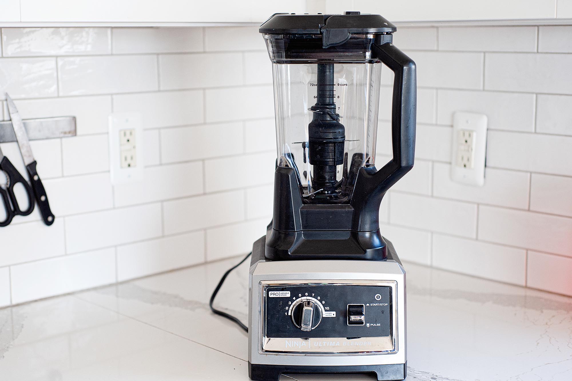 An empty high speed blender with base and lid are on a white countertop. White subway tile backsplash and knife magnetic strip with a knife and scissors is to the left.