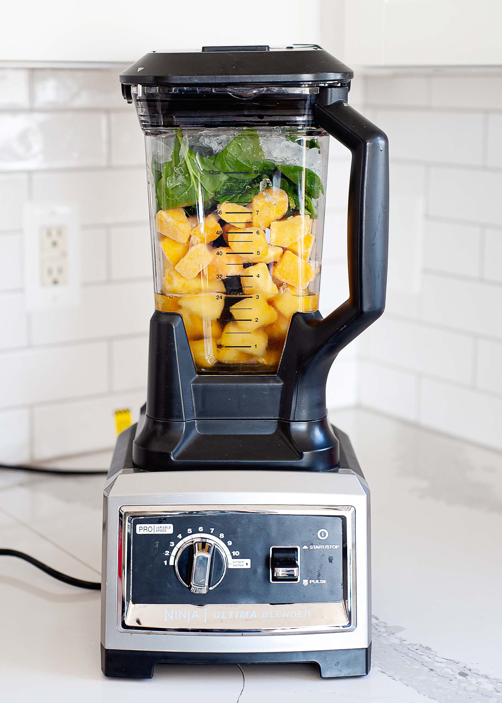 View of a high speed blender with pineapple and spinach inside. The blender sits on a white counter with subway tiled backsplash behind it.