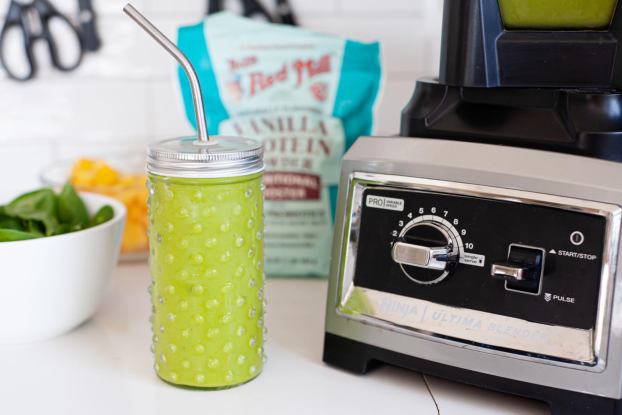 A green smoothie in a lidded glass mason jar with a stainless steel straw sits on a white countertop. The base of a high speed blender is to the right of the smoothie. A bowl of spinach, cut fruit and a bag of protein powder are to the left of the smoothie as well as behind it.