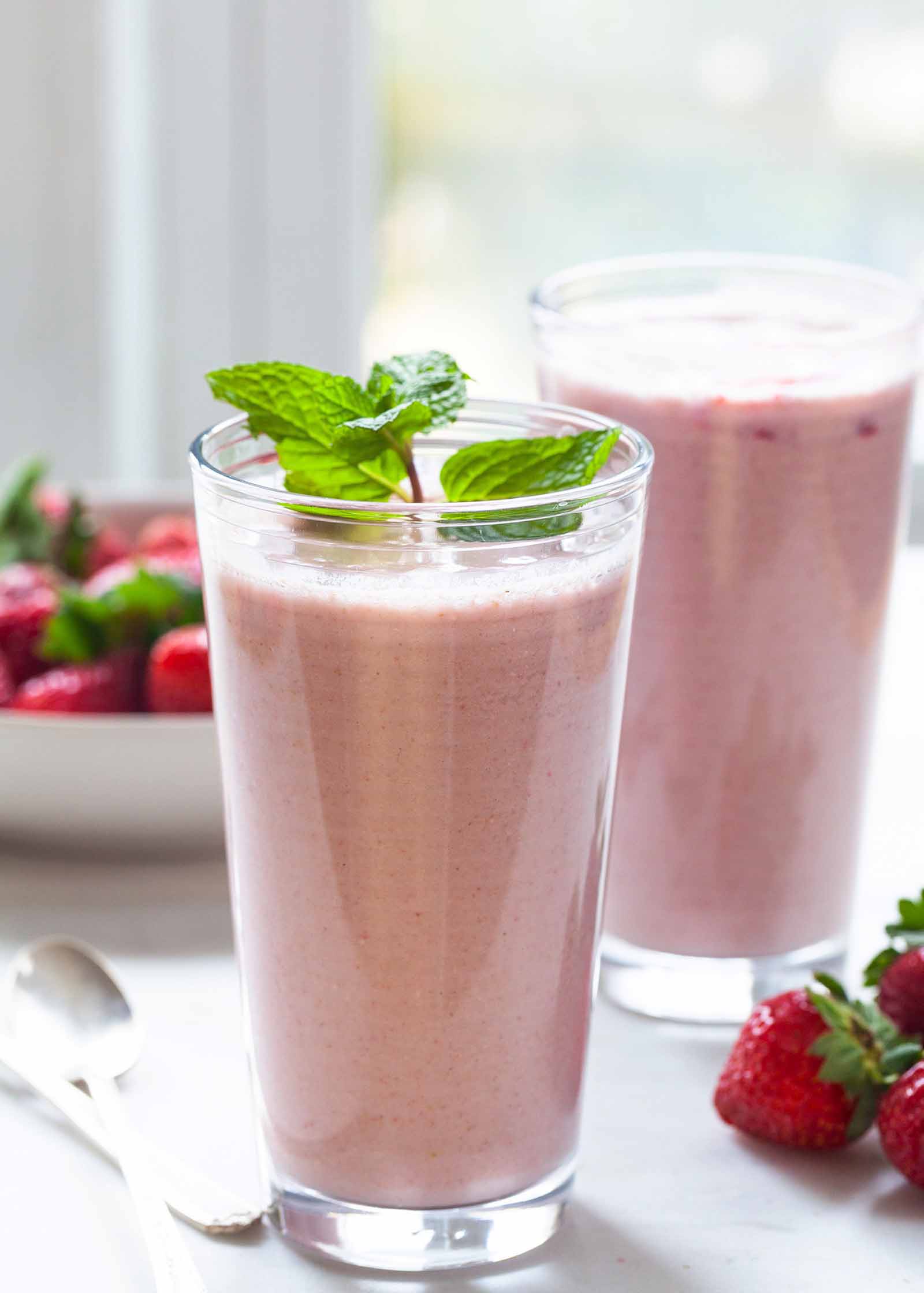 Side view of two tall glasses of healthy strawberry almond oat smoothie. Whole strawberries are on the table to the right of the glass in front and two spoons are to the left. A bowl of whole strawberries are behind the glasses.