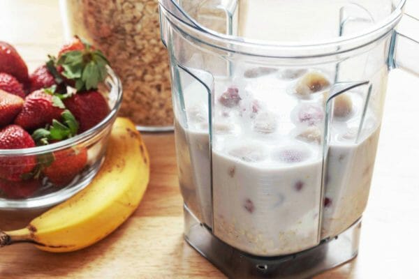 A blender of easy strawberry almond oat smoothie ingredients. A slightly brown banana, bowl of strawberries and container of oats are to the left of the blender.