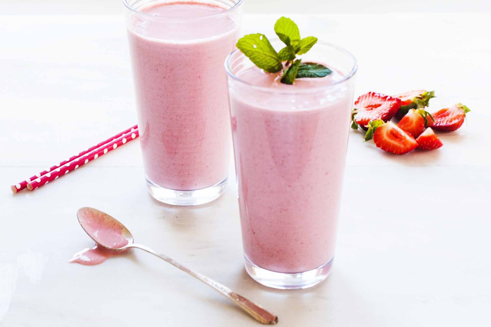 Two tall breakfast smoothies in tall glasses. One has a sprig of mint on top. Clockwise around the glasses are sliced strawberries, a spoon with the pink smoothie on it. Lastly, are two bright pink straws behind the glass.