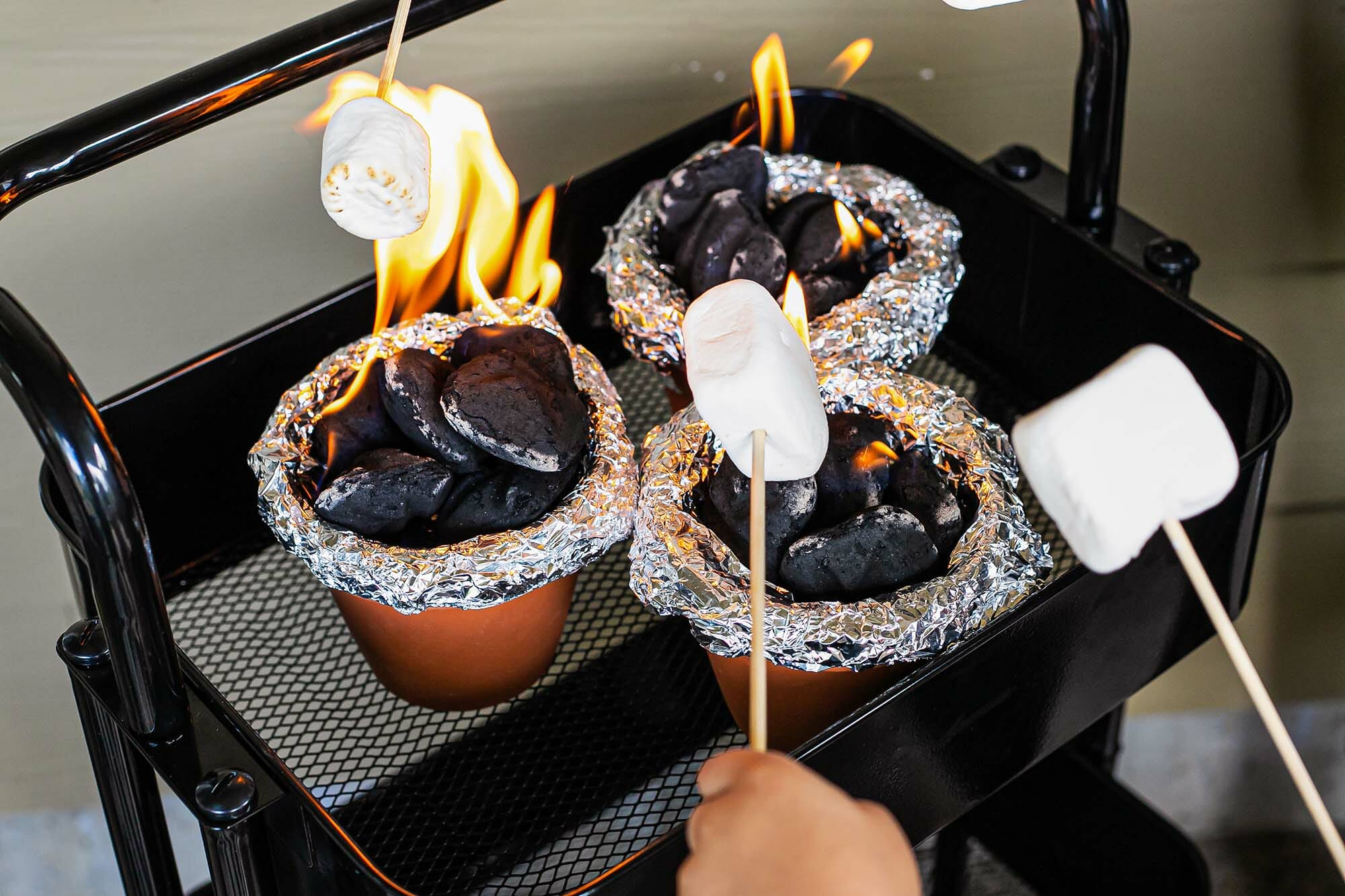 Marshmallows are roasting over a terra cotta pot with coals inside to show how to make a smores.