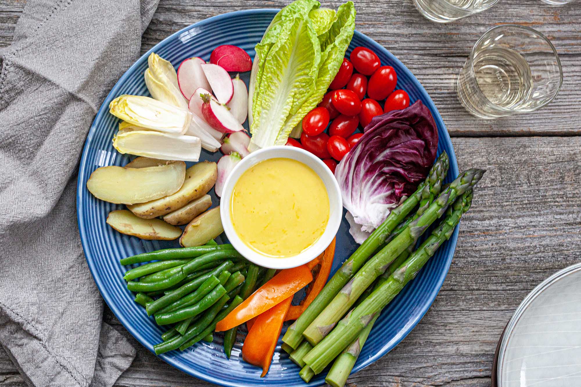 A plate with homemade aioli in a small bowl in the center of a plate. The aioli sauce is surrounded by a variety of steamed and fresh vegetables.