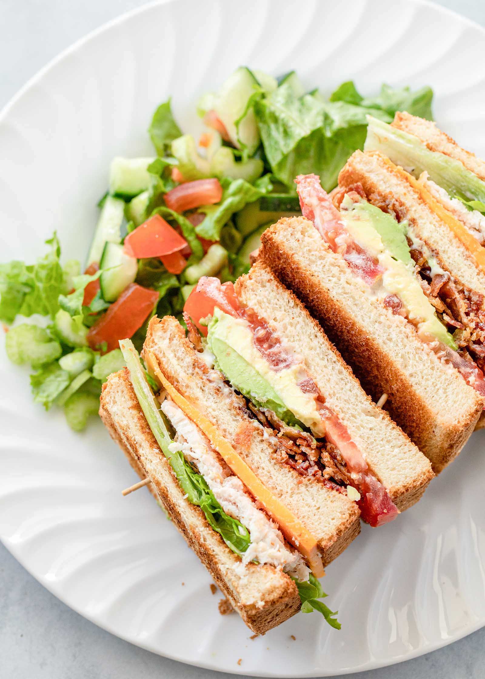 Overhead view of a turkey club sandwich on its side. A lettuce salad with cucumber and tomato is on the plate behind the sandwich.