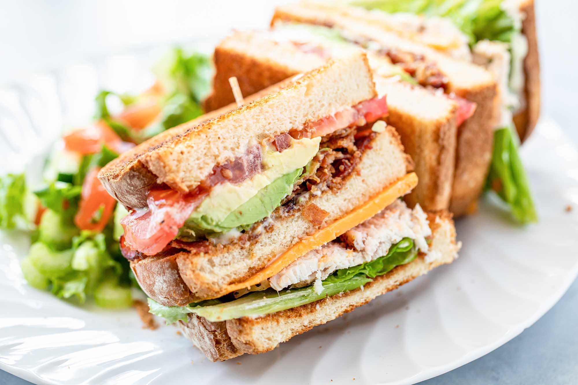 Side view of turkey club with garlic mayo on a plate. The bread is toasted and the mayo, avocado, bacon, tomato, lettuce and turkey are visible in the sandwich.