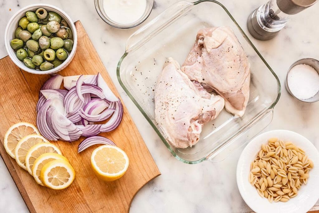 Marble countertop with ingredients for yogurt marinated lemon chicken with pasta, olives and herbs. A wooden cutting board to the left of the photo has sliced lemons, sliced red onions and a small bowl of green olives on it. To the right of the cutting board is a glass casserole dish with two bone-in chicken breasts. Below the casserole dish is a small white bowl of shell pasta. Salt an pepper are int he upper left corner of the photo.