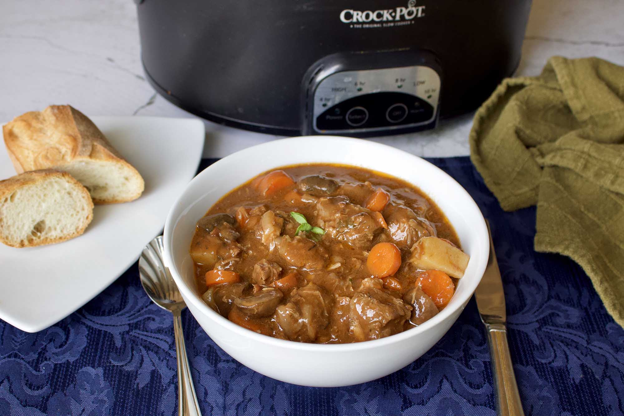 Slow Cooker Beef Burgundy (Boeuf Bourguignon) in a bowl set in front of a crock pot. Silverware, bread, a place mat and kitchen towel are surrounding the bowl.