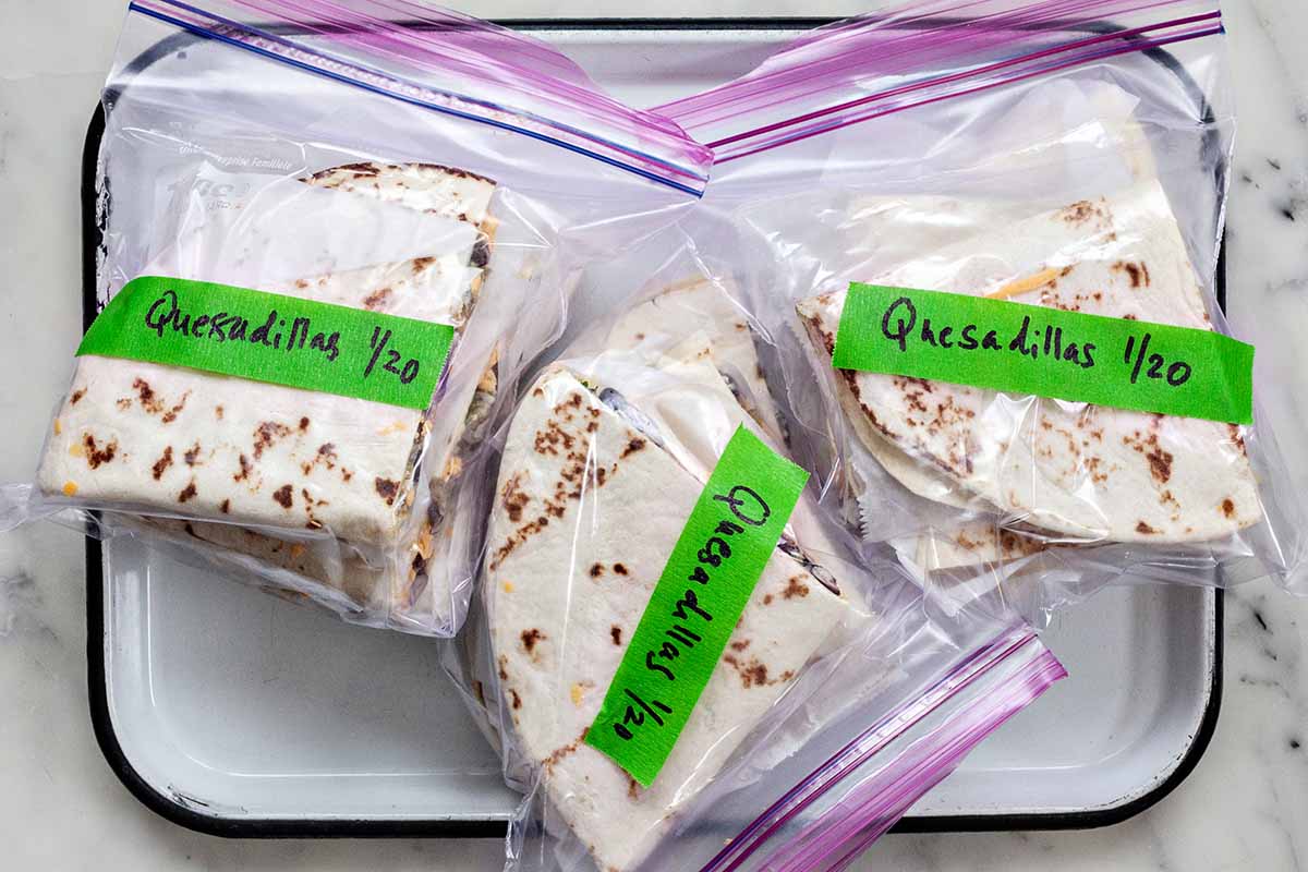 Overhead view of three ziplock freezer bags with simple breakfast quesadilla triangles inside. Each bag is labeled "Quesadillas 1/20" with green painter tape. The ziplocks are on a white tray on a marble countertop.