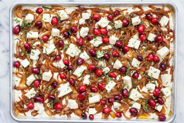 Foccaccia in a sheet pan and topped with caramelized onion, cranberry, and brie and ready to be baked.