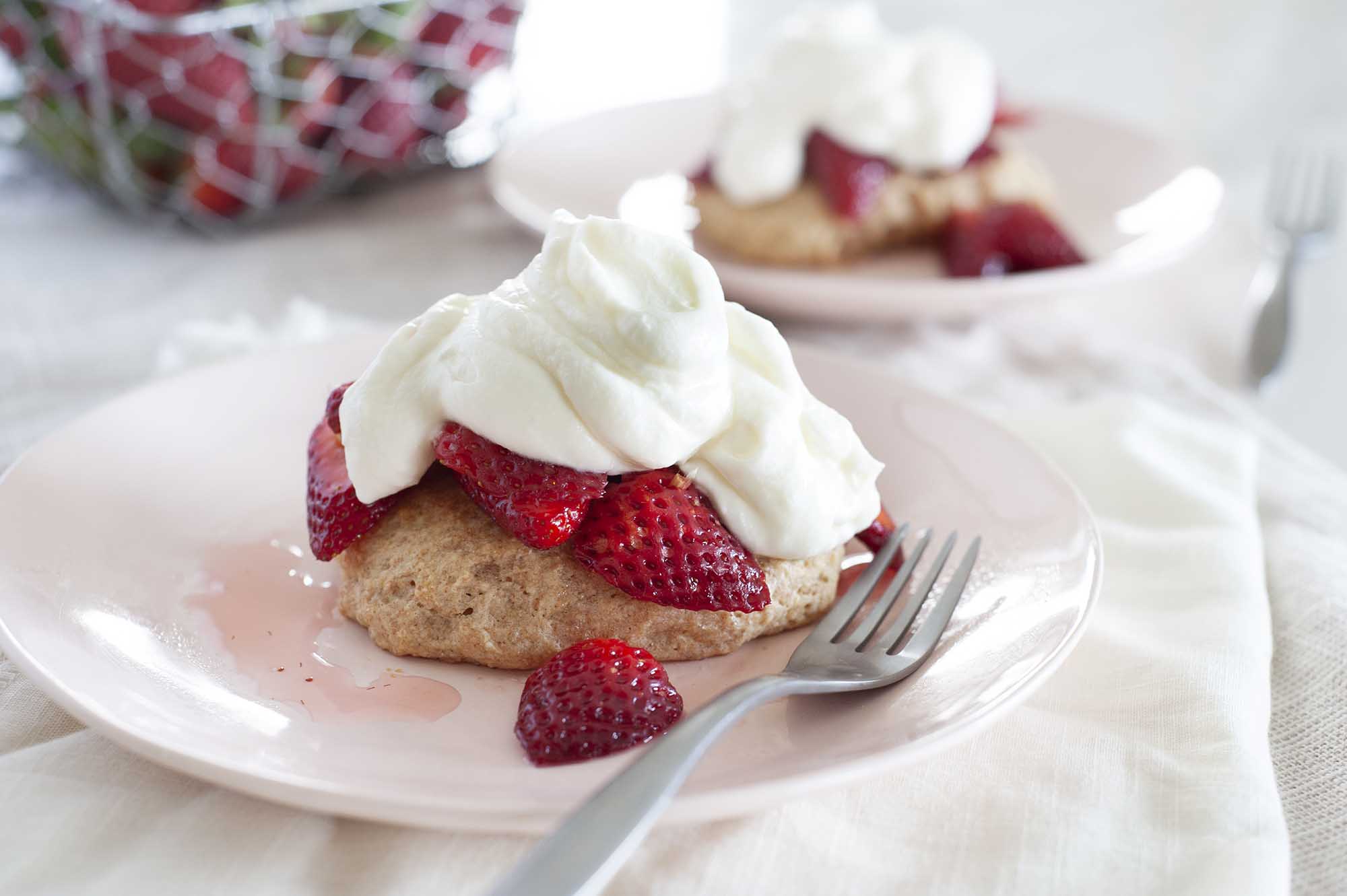 Two plates with easy strawberry shortcake set on a table with white linens. Shortcake is piled with sliced strawberries and whipped yogurt topping. A wire basket of strawberries is to the left of the plates.